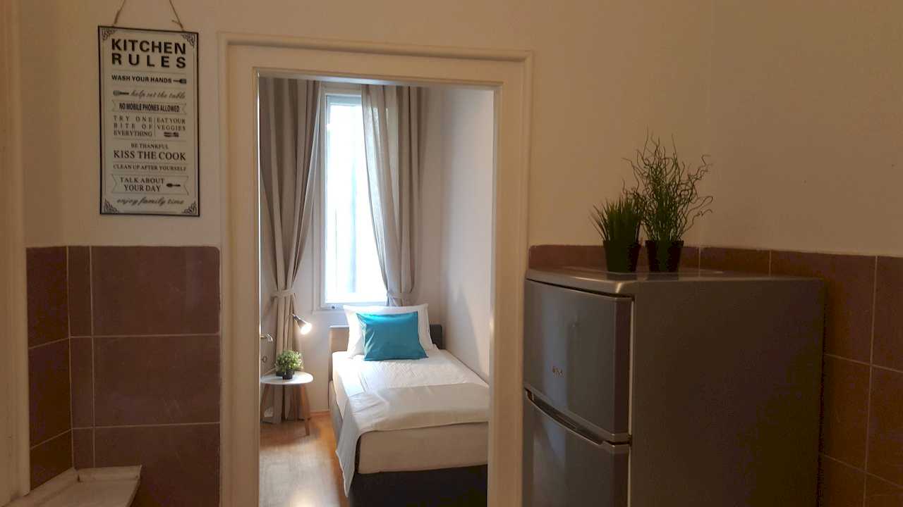 View from kitchen through doorway into narrow bedroom with single bed; fridge with plants and Kitchen Rules wall art – Apartment Opera – City Break Apartments