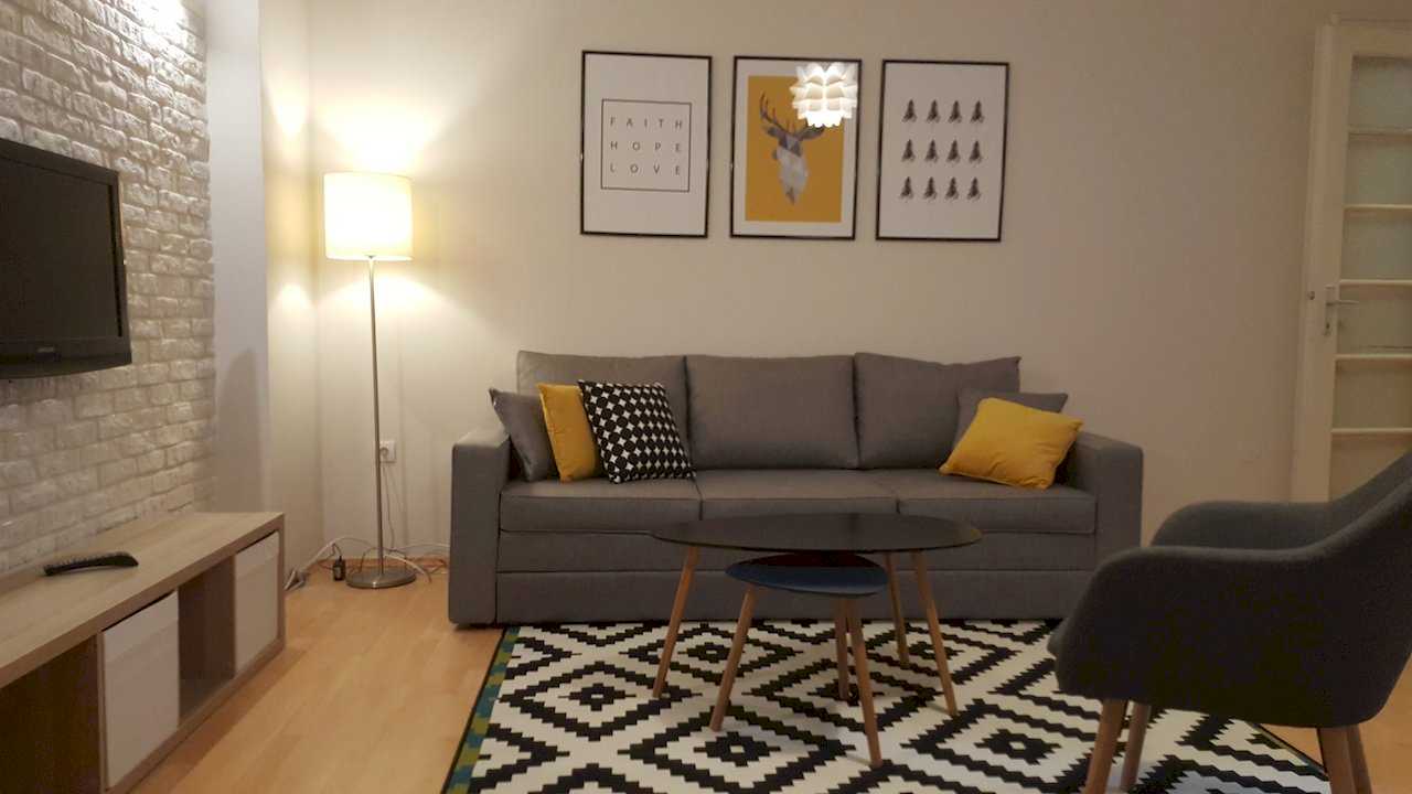 Living room with grey sofa, yellow cushions, three framed prints, modern pendant, coffee table on rug, and TV on white brick wall – Apartment Opera – City Break Apartments