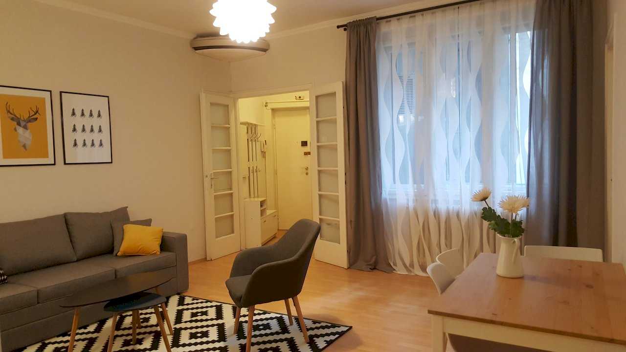 Open-plan living and dining with sofa, armchair, coffee table, dining table with white flowers, curtains, and entry hallway view – Apartment Opera – City Break Apartments