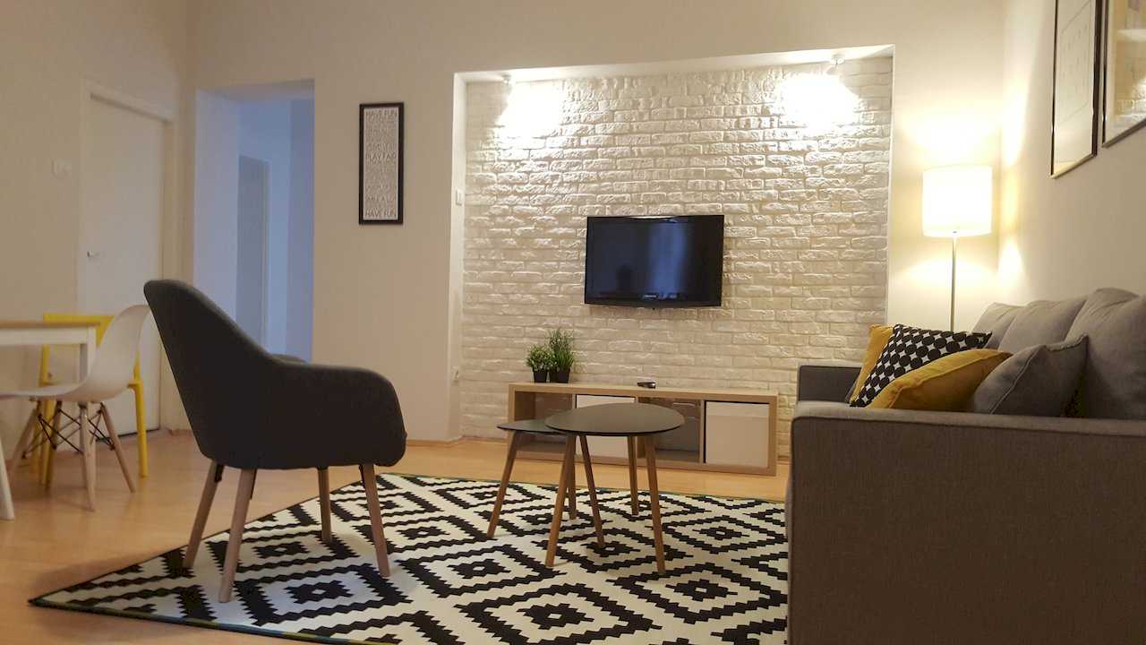 Living area with white brick TV niche and spotlights, grey sofa with yellow pillows, armchair, and twin round coffee tables – Apartment Opera – City Break Apartments