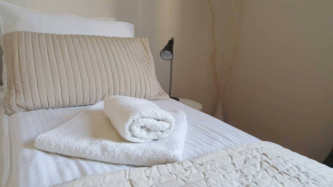 Close-up of double bed with striped white bedding, beige pillow, folded white towels, bedside lamp, and a vase with decorative branches in the background – Apartment Opera – City Break Apartments