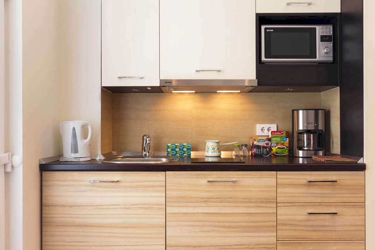 Compact kitchen with white upper and wood-grain lower cabinets, built-in microwave, hood, kettle and coffee maker on a dark worktop – Apartment Trg 1 – City Break Apartments