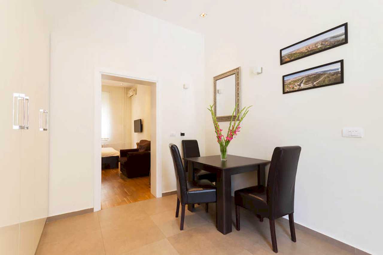Dining area with dark wood table, black chairs, vase of pink flowers, panoramic prints and wall mirror, view toward the living zone – Apartment Trg 1 – City Break Apartments