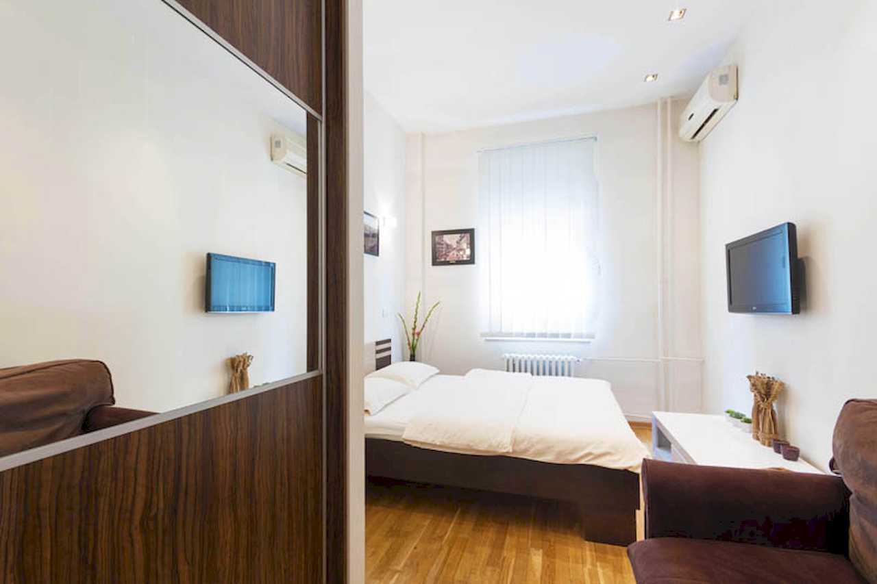 Bedroom with double bed, wall-mounted TV and air conditioner, mirrored sliding wardrobe and brown armchair – Apartment Trg 1 – City Break Apartments