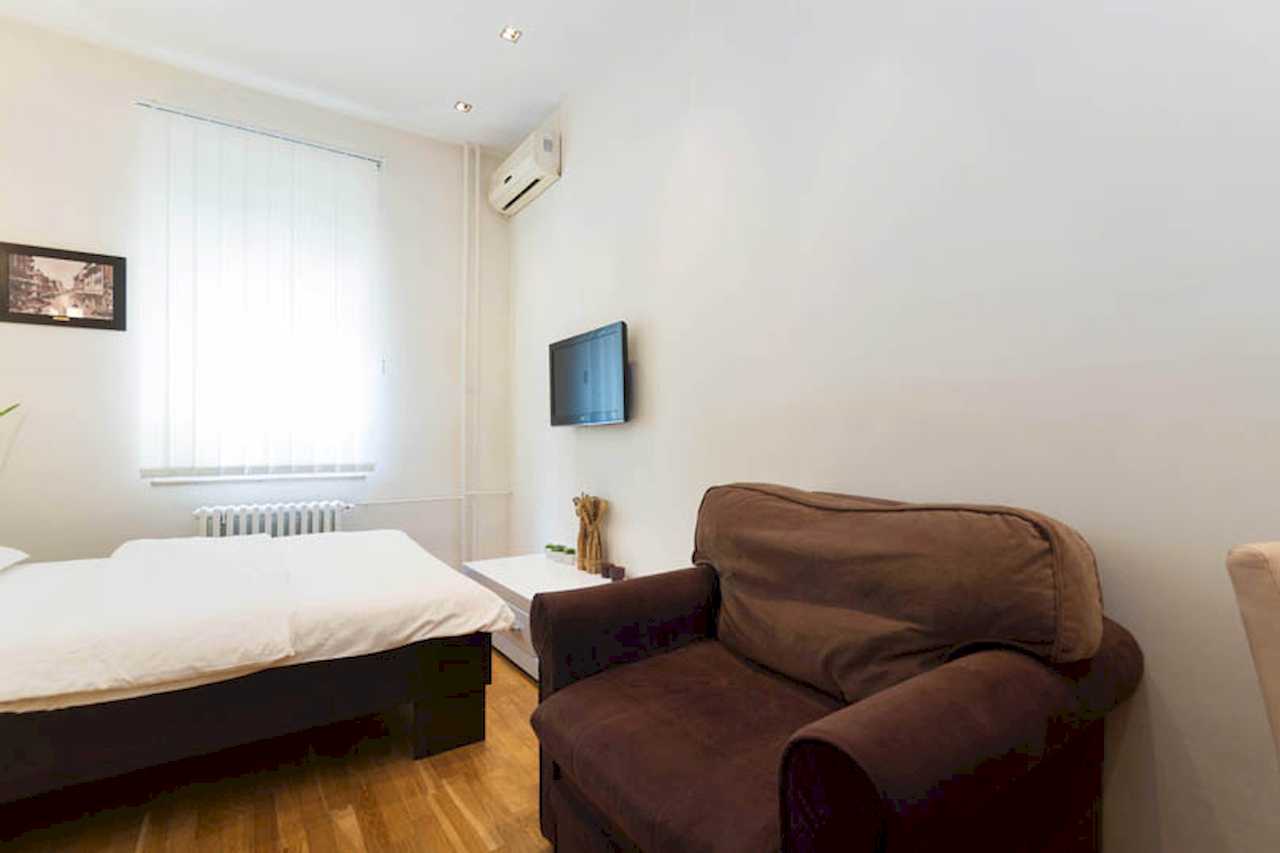 Bedroom with double bed, brown armchair, wall-mounted TV and air conditioner, window with white vertical blinds – Apartment Trg 1 – City Break Apartments