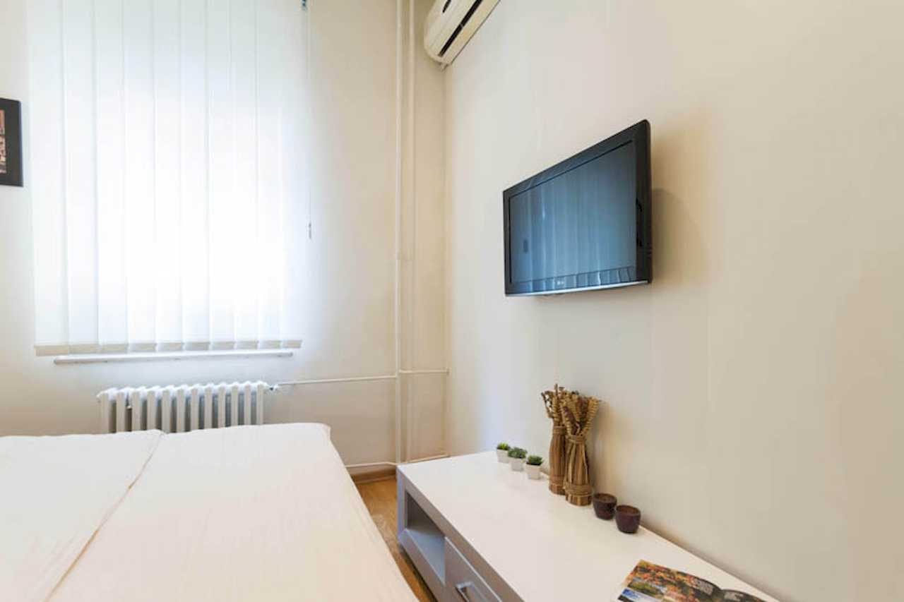 Wall-mounted TV above white dresser with plants and decorative vases, vertical blinds and radiator in the bedroom – Apartment Trg 1 – City Break Apartments
