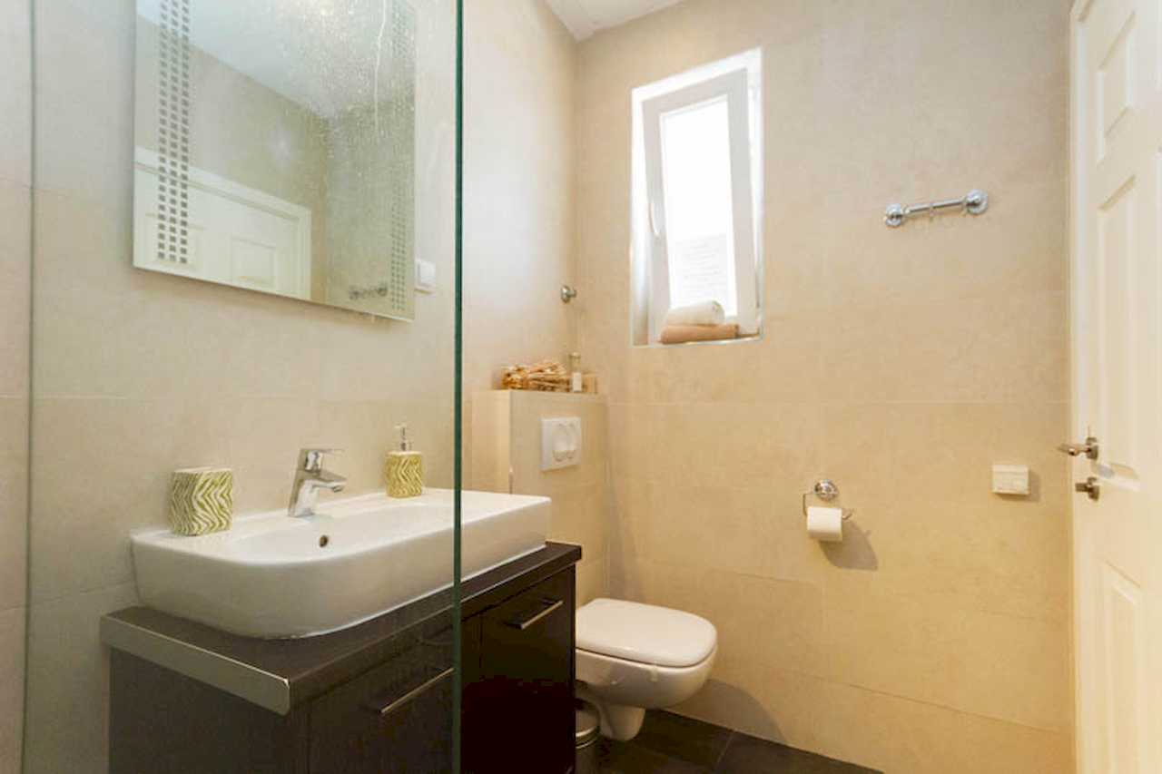 Bathroom with white wall-mounted sink on dark floating vanity, wall-hung toilet, narrow window and glass shower screen – Apartment Trg 1 – City Break Apartments