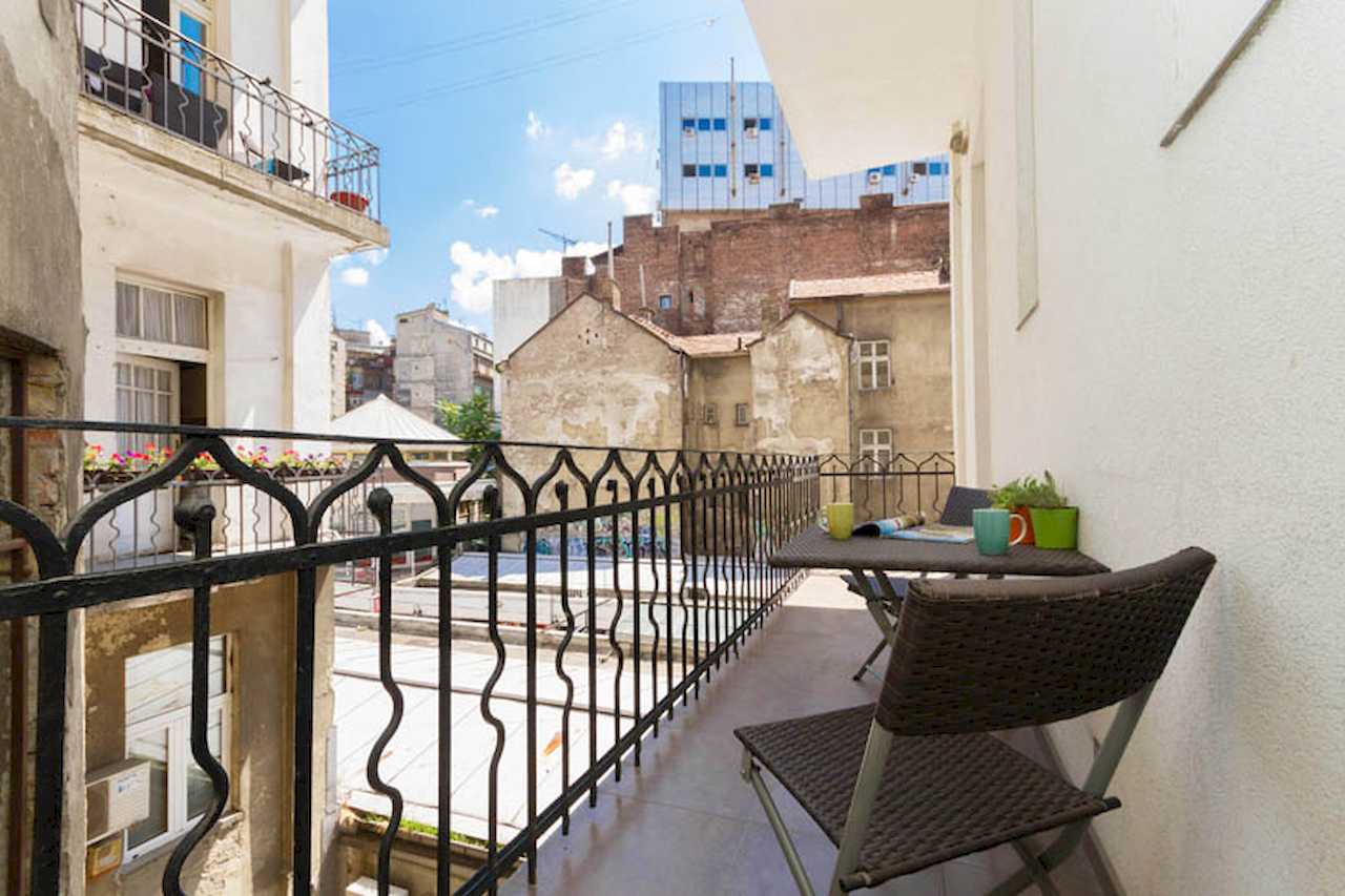 Balcony with dark outdoor table and two chairs, mugs and plants on the table, wrought-iron railing and view into the inner courtyard – Apartment Trg 1 – City Break Apartments