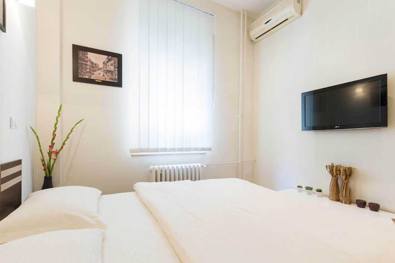 Bedroom with double bed, wall-mounted TV and air conditioner, vase of gladioli, framed print and white shelf under the TV – Apartment Trg 1 – City Break Apartments