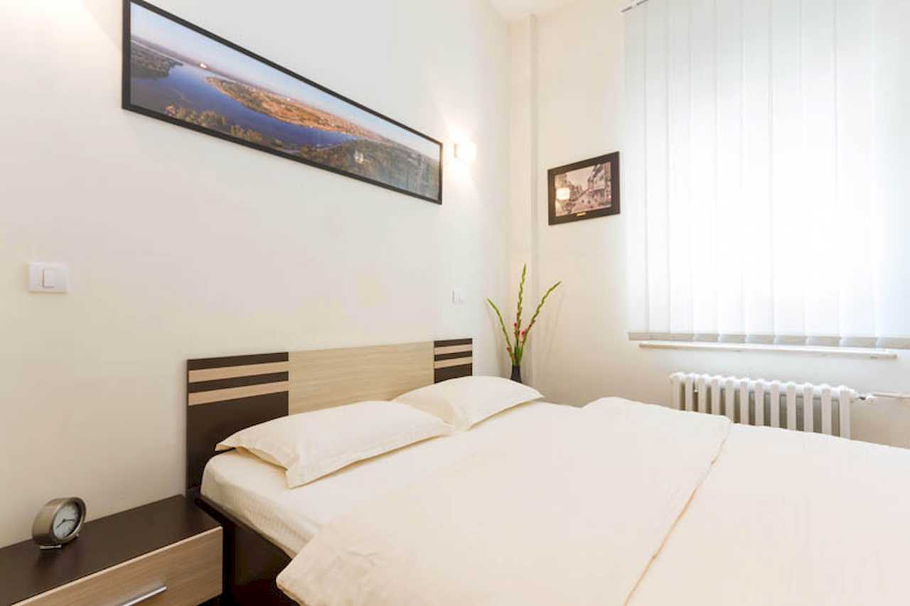 Bedroom with double bed, panoramic river-city print above the bed, vertical blinds and bedside table with clock – Apartment Trg 1 – City Break Apartments