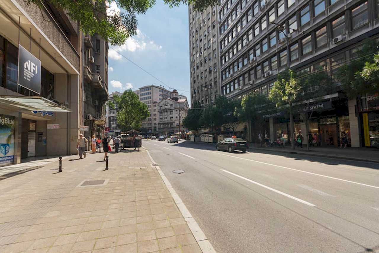 View of a wide central street: tree-lined pavement, ground-floor shops, traffic and pedestrians – Apartment Trg 1 – City Break Apartments