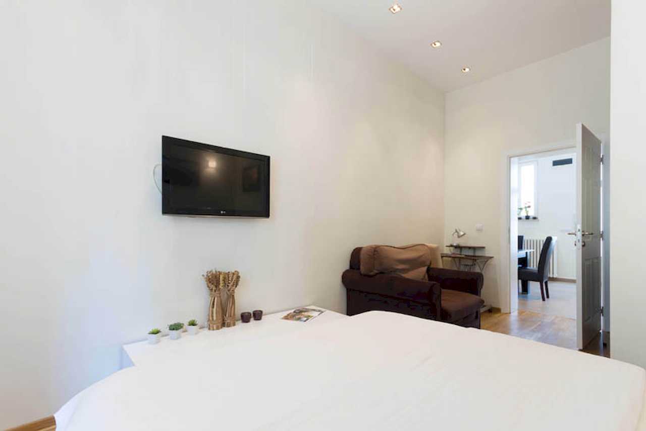 Bedroom with double bed, decorative accents on the headboard wall, wall TV, brown armchair and desk, view toward the dining area – Apartment Trg 1 – City Break Apartments