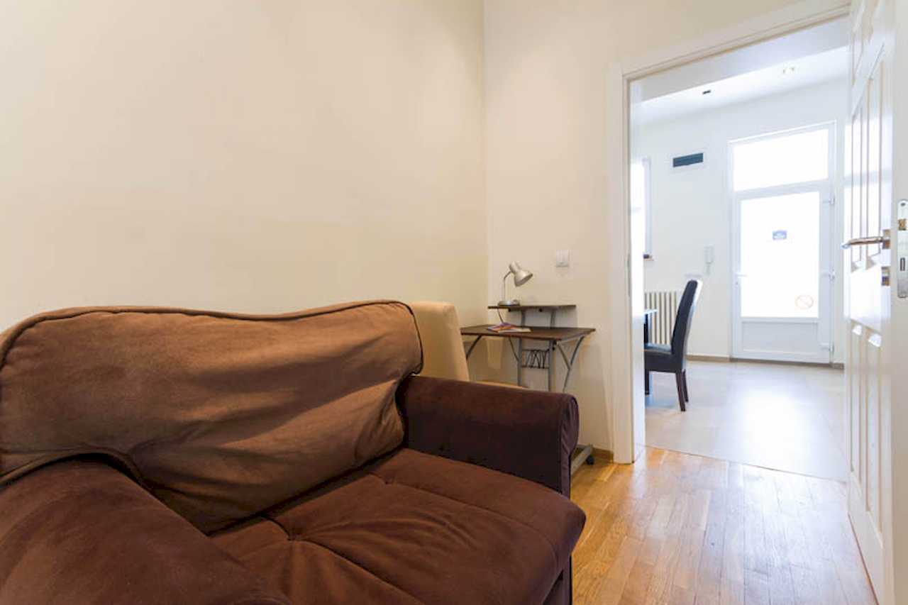 Room corner with brown armchair, small desk with lamp and view through open doors to the dining area and entrance – Apartment Trg 1 – City Break Apartments