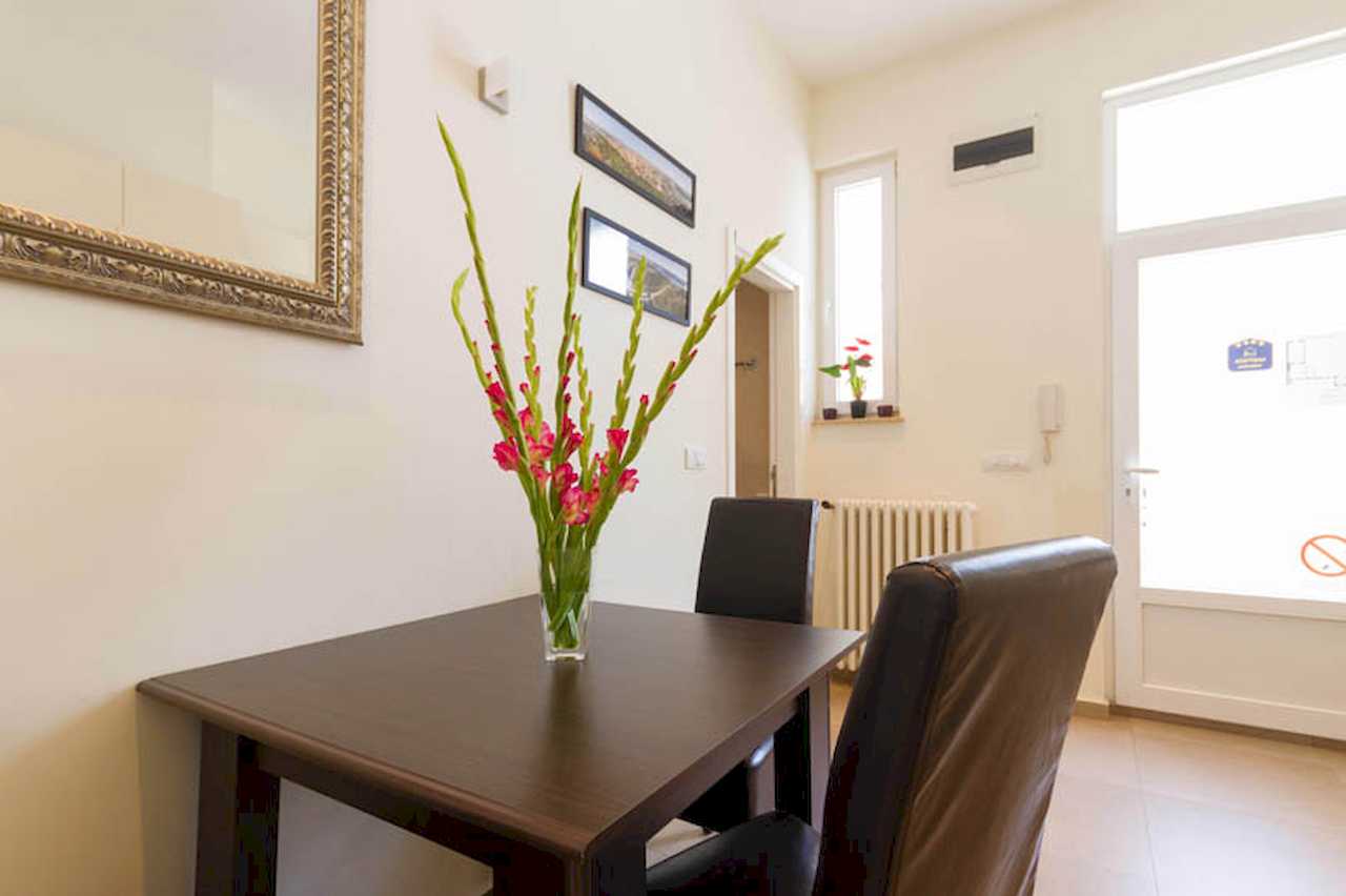 Dining table with pink flowers in a vase, gold-framed mirror, panoramic prints on the wall and glazed entrance door – Apartment Trg 1 – City Break Apartments