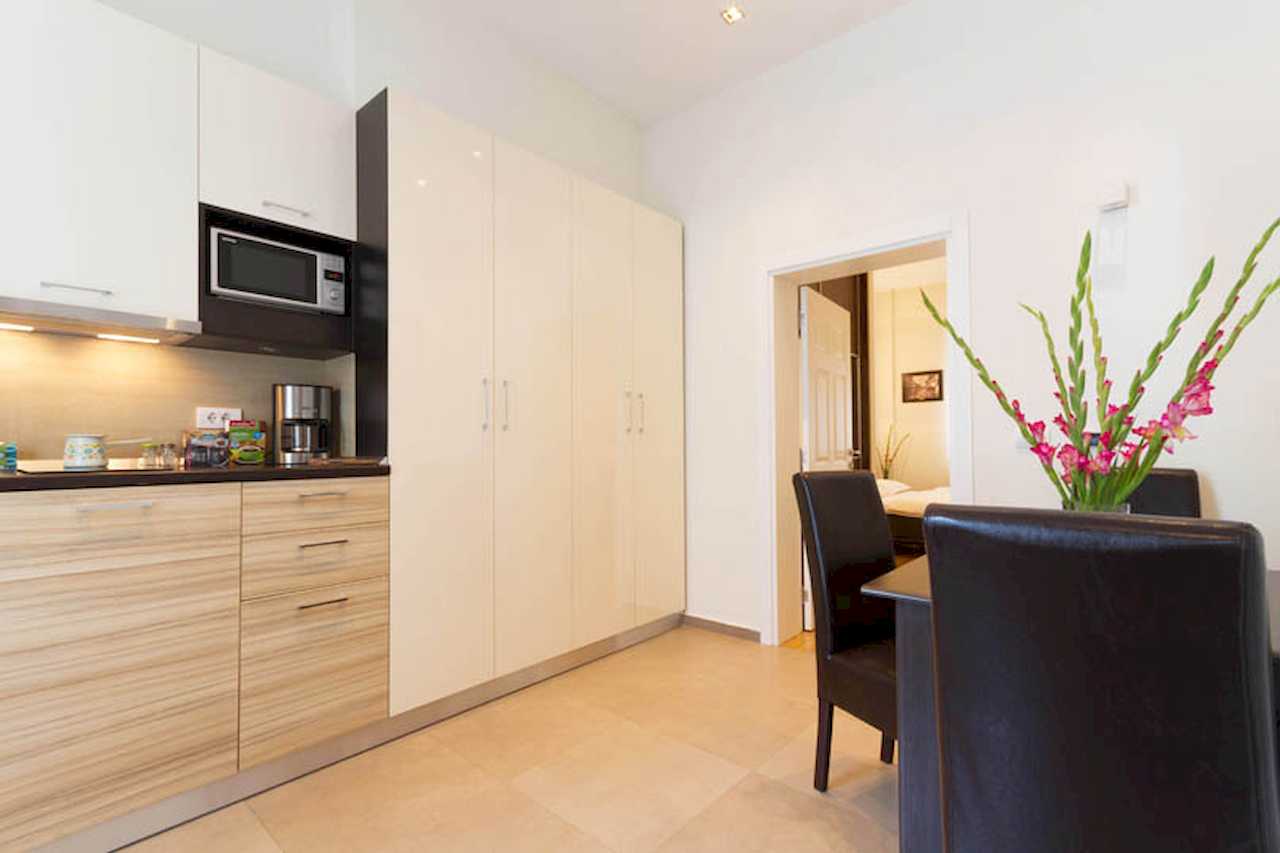 Kitchen with white and wood fronts, tall white wardrobe, dining table with flowers and open door to the bedroom – Apartment Trg 1 – City Break Apartments
