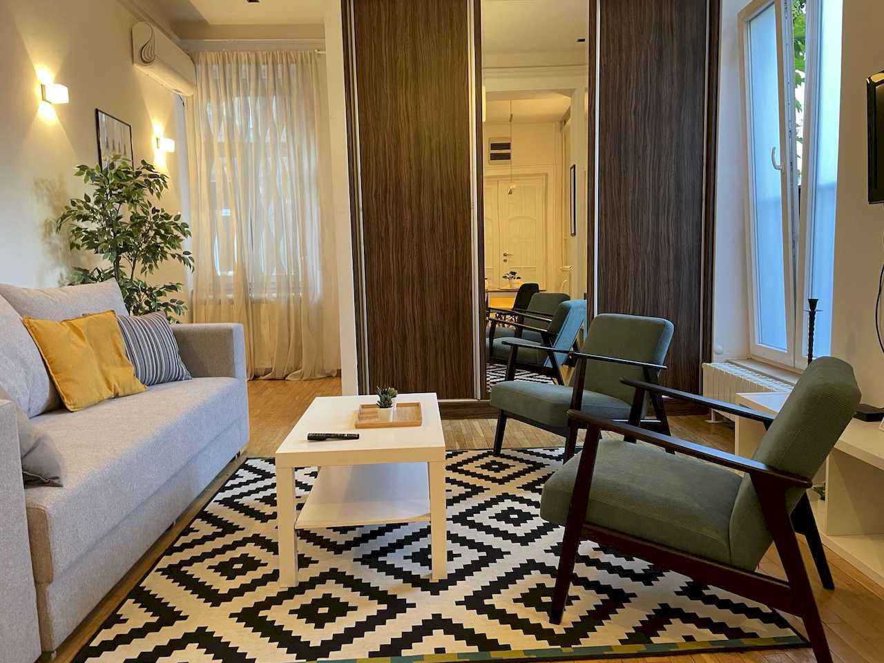 Dnevna soba sa sivom sofom, dve tirkizne fotelje i veliki plakar sa ogledalom – Apartman Trg 2 – City Break Apartments