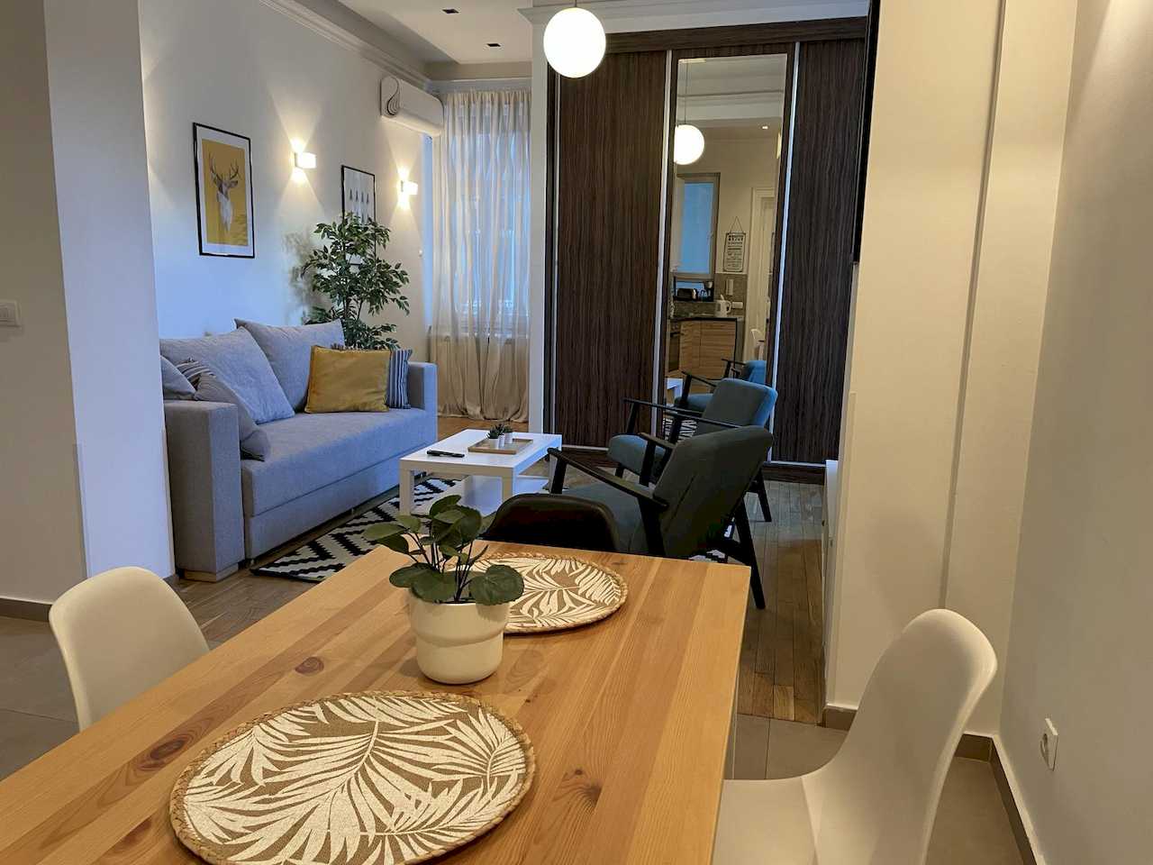 Trpezarijski sto i dnevna soba sa sofom, foteljama i belom kugla visilicom – Apartman Trg 2 – City Break Apartments