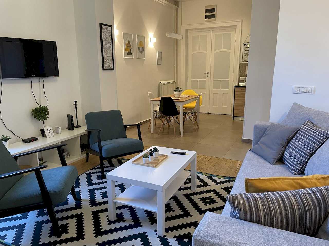 Dnevna soba sa TV-om, sofom, foteljama i trpezarijom u pozadini – Apartman Trg 2 – City Break Apartments