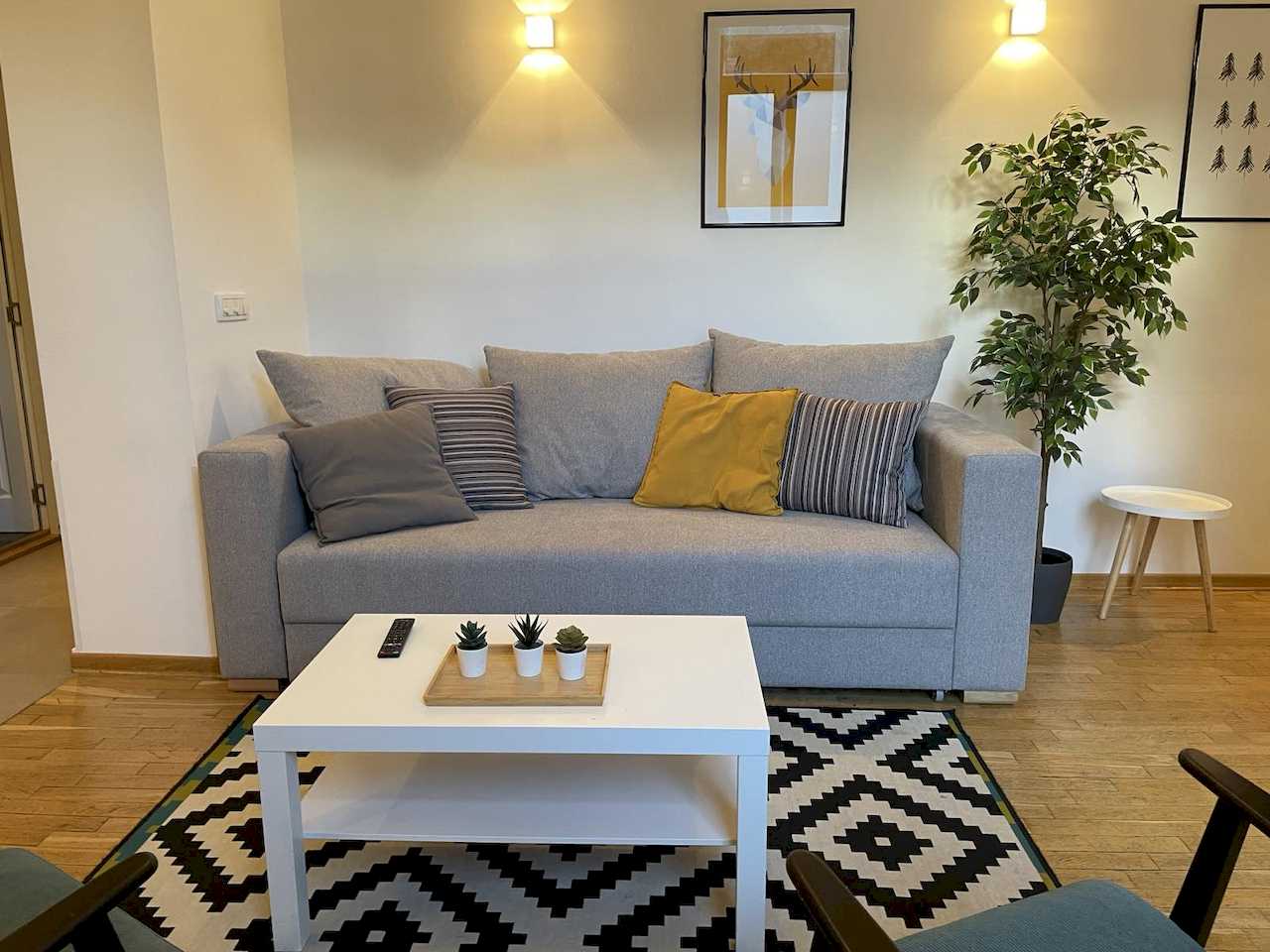 Siva sofa sa žutim jastukom, beli klub sto i geometrijski crno-beli tepih – Apartman Trg 2 – City Break Apartments