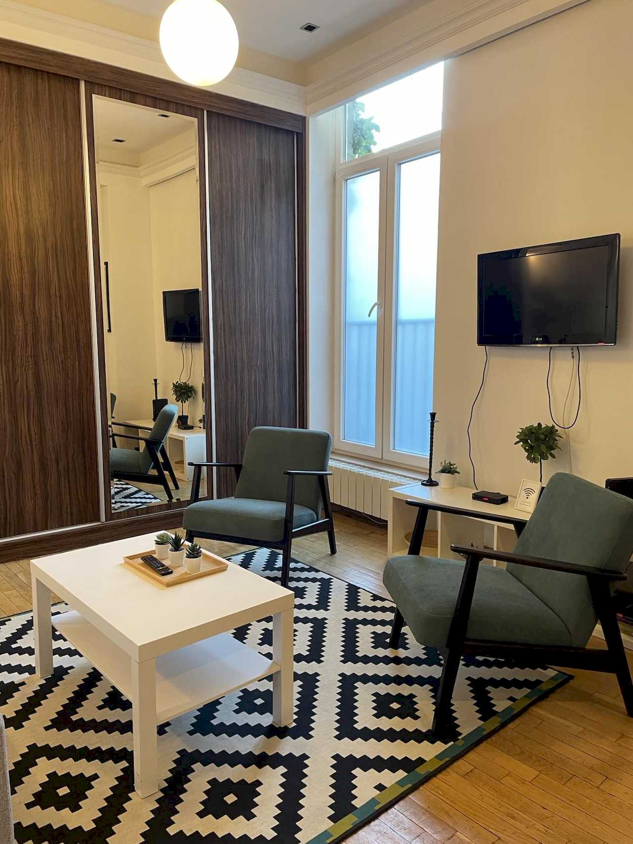 Dve fotelje, beli klub sto i plakar sa ogledalom ispod zidnog TV-a – Apartman Trg 2 – City Break Apartments
