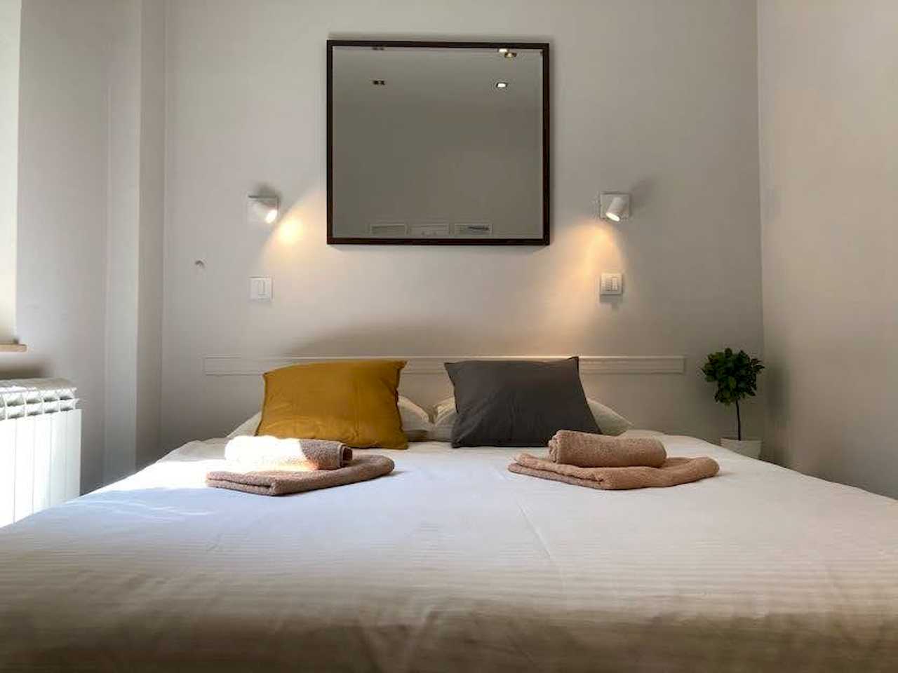 Spavaća soba sa bračnim krevetom, kvadratnim ogledalom i uključenim zidnim lampama – Apartman Trg 2 – City Break Apartments