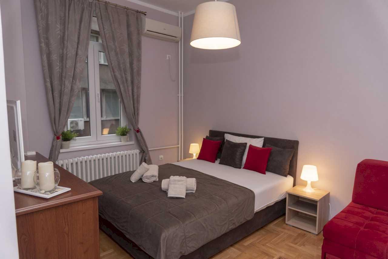 Četvorosobni stan Trg 4 (Kolarčeva, Beograd) – spavaća soba sa bračnim krevetom, sivim i crvenim jastucima i prozorom sa zavesama